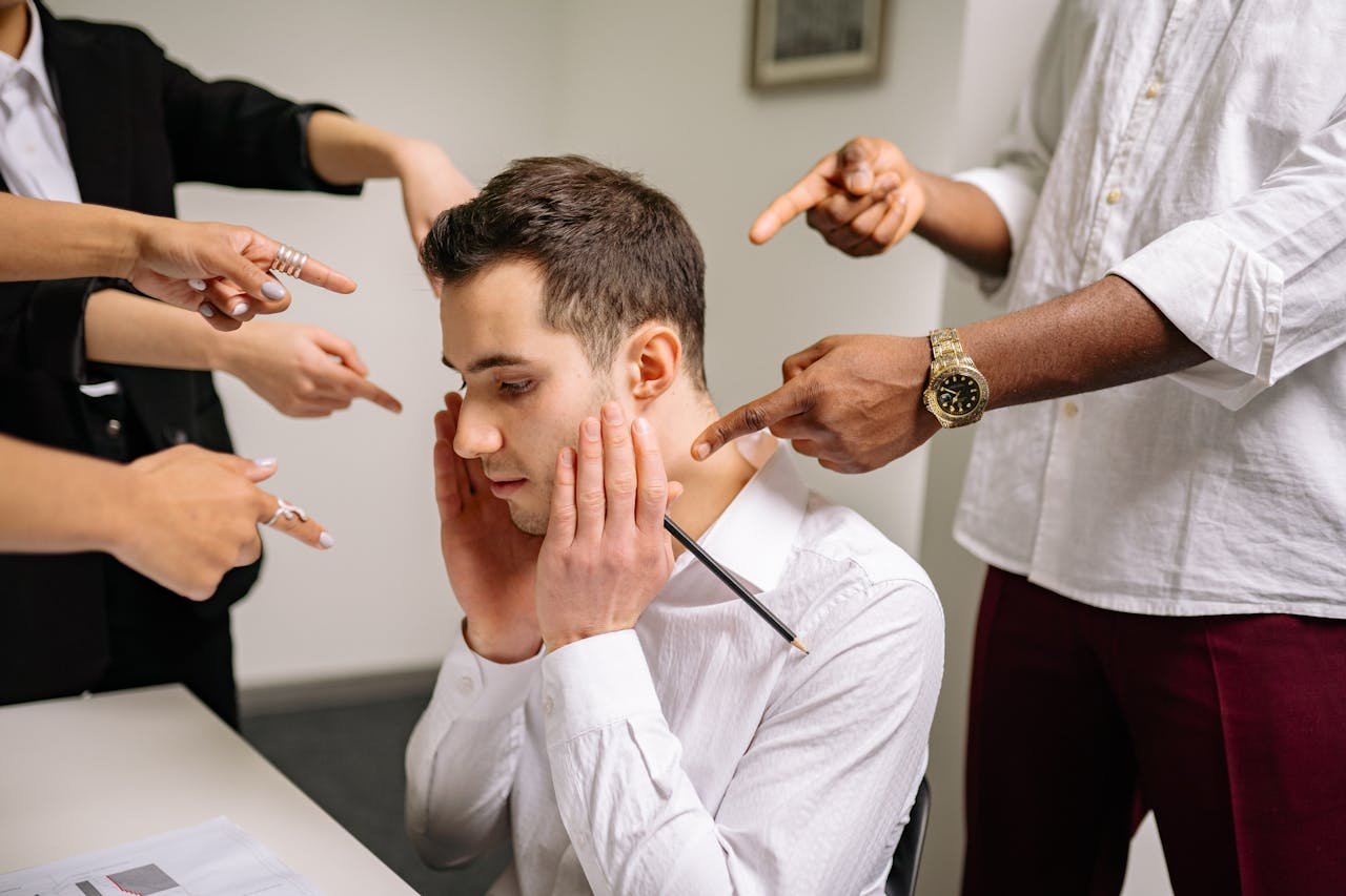 A stressed man at the office surrounded by pointing fingers, representing workplace bullying and stress.